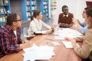 Equipe de jovens discutindo ideias em mesa de biblioteca com laptop e documentos, em cenário de escolha da melhor franquia de escola