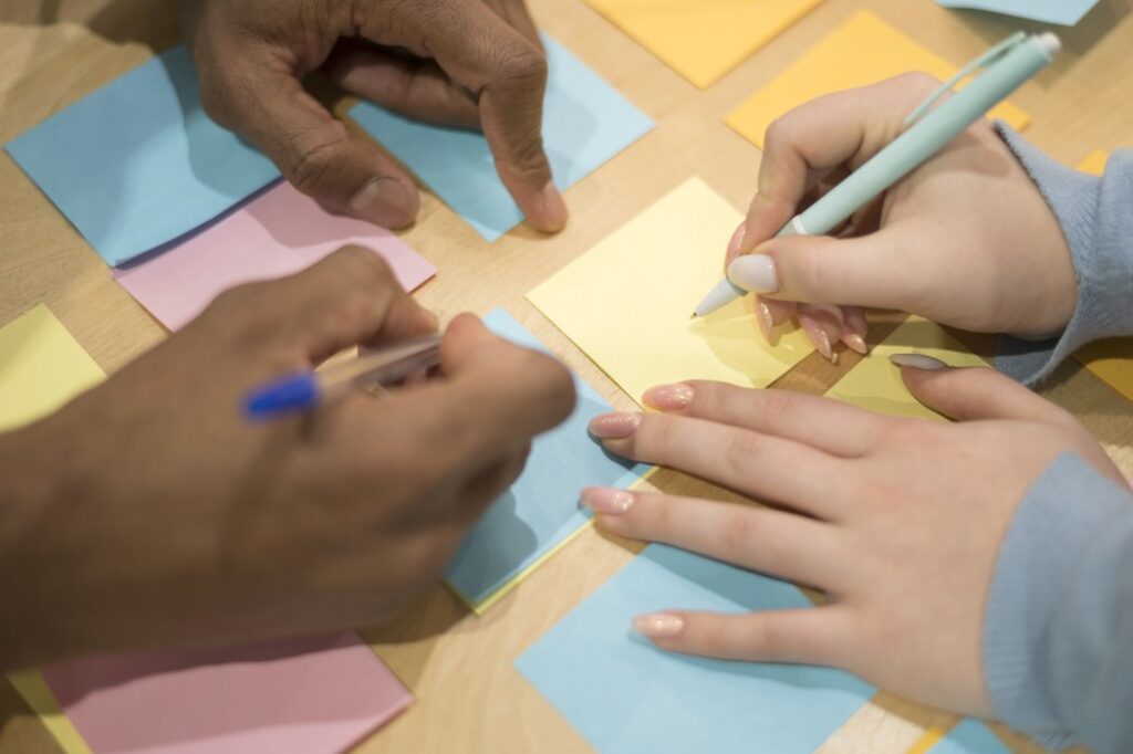 Mãos de pessoas de diferentes origens escrevendo em post-its coloridos durante atividade colaborativa, representando a troca cultural na educação cristã intercultural