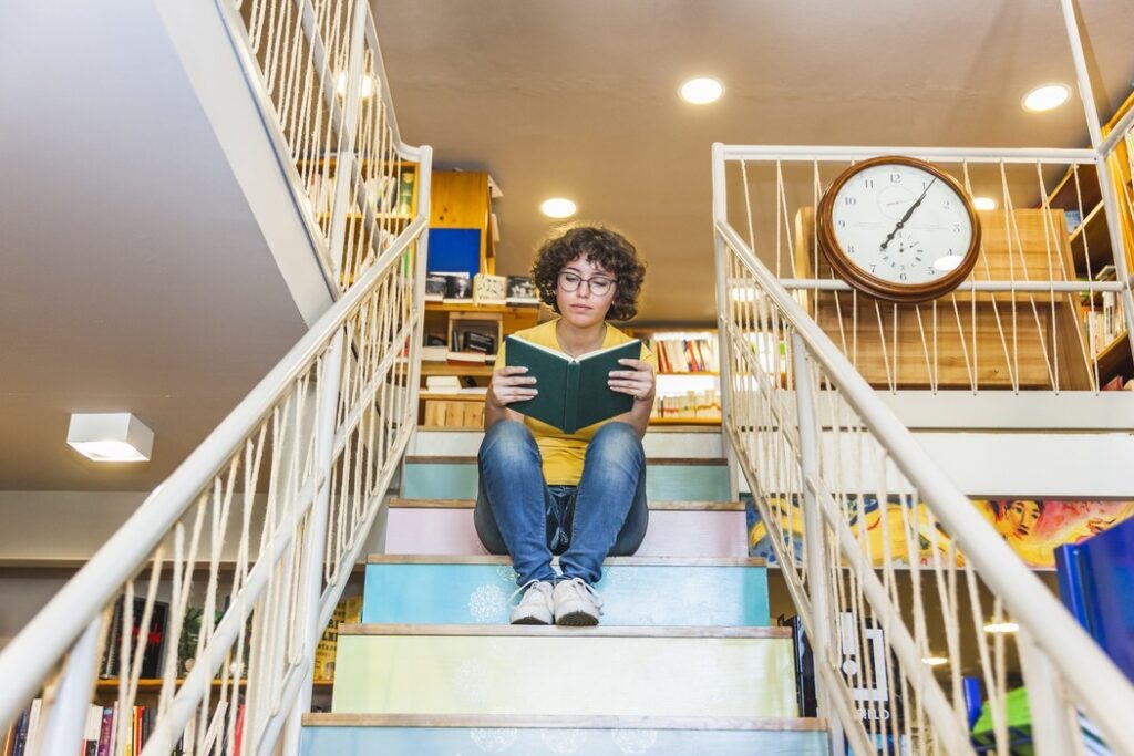 Estudante lendo livro sentada em escada colorida de biblioteca, representando ambiente de aprendizagem valorizado em franquia de escola particular
