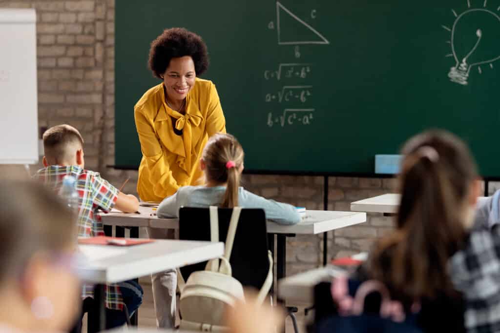 Professora sorridente de blusa amarela inclinada para conversar com aluno em sala de aula, com lousa verde com fórmulas matemáticas ao fundo, ilustrando conteúdo sobre mercado educacional